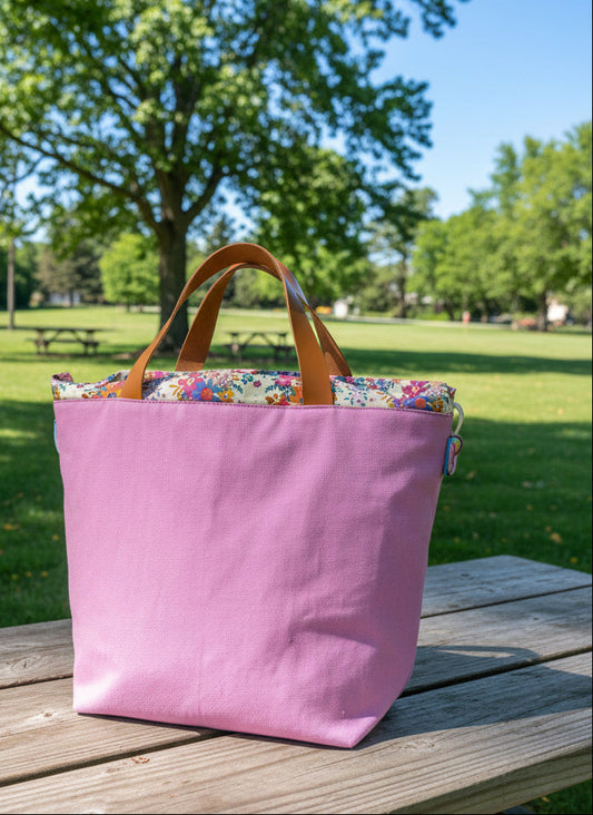 A lavender canvas tote bag with brown leather handles and a colorful floral-patterned lining, shown against a draped white fabric background.