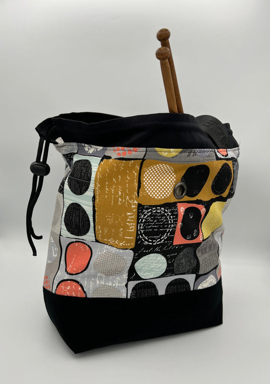 A handmade knitting bag with a black bottom and a colorful abstract patchwork pattern on the upper half. A wooden double-pointed knitting needle rests inside, and a black drawstring is visible on the left side.