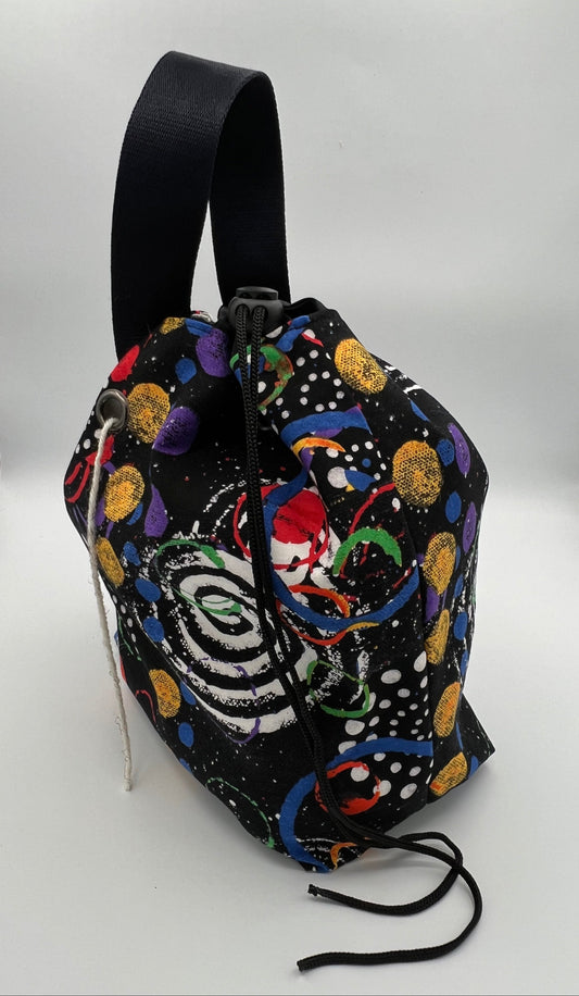 A black drawstring bag with a colorful cosmic print featuring planets, stars, and a spiral galaxy design, complete with a wide black strap, set against a white studio background.