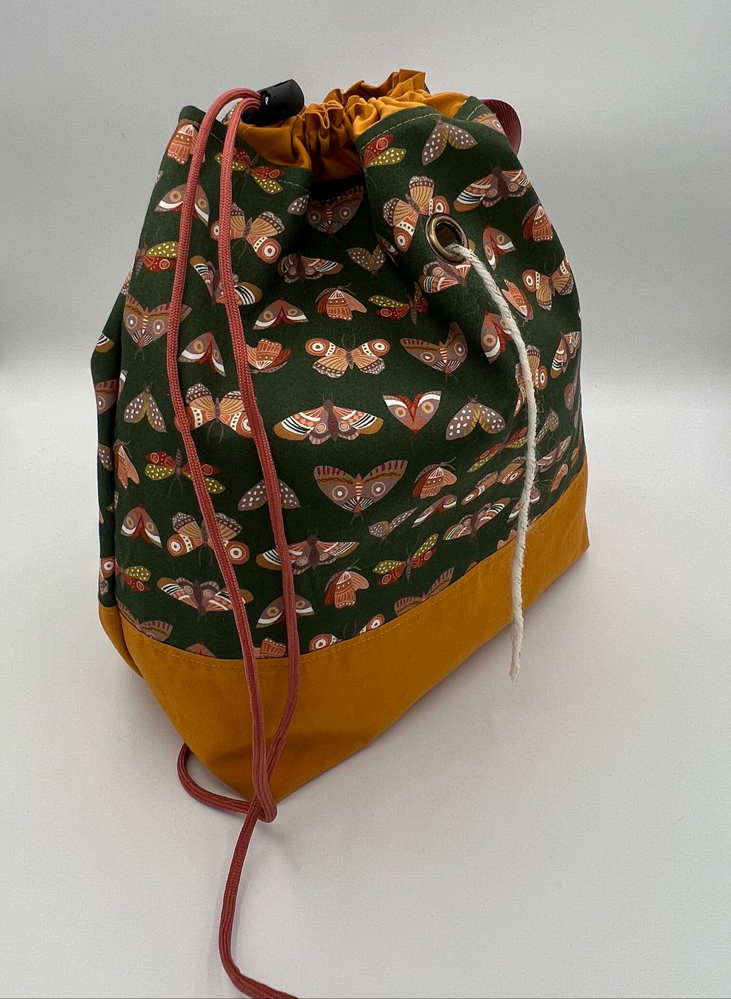 A fabric drawstring bag with a dark green upper section featuring a butterfly pattern, a mustard yellow bottom, and rust drawstrings, displayed on a white studio background.