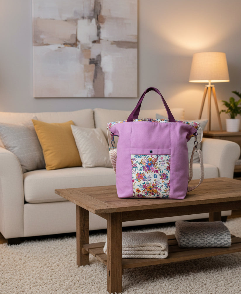 A lavender canvas tote bag with a floral print front pocket and matching floral lining, featuring dark purple straps and a beige rope-style strap detail, set against a draped white fabric background.