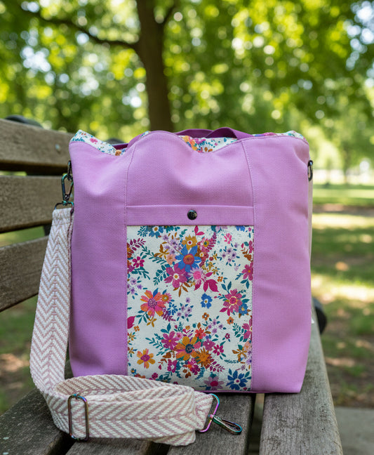 A lavender canvas shoulder bag with a colorful floral patterned front pocket and a beige woven strap accessory, shown on a white background.