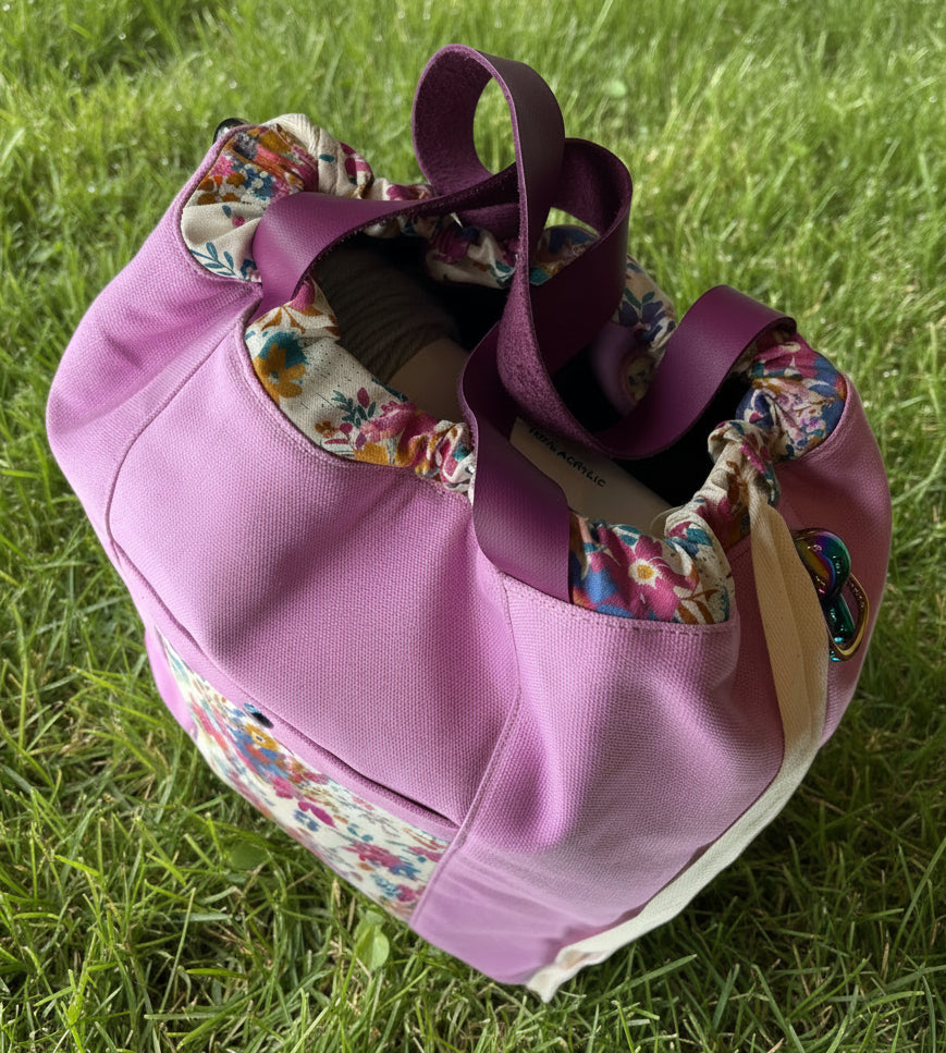 A lavender canvas tote bag with floral patterned fabric trim and dark purple shoulder straps with drawstring cinch.  