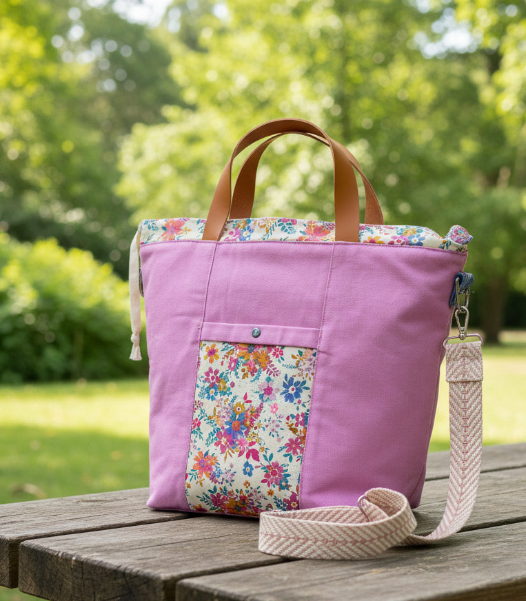 A lavender canvas tote bag with a colorful floral patterned front pocket and top trim, featuring brown leather top handles and a beige woven shoulder strap, displayed on a white fabric surface.