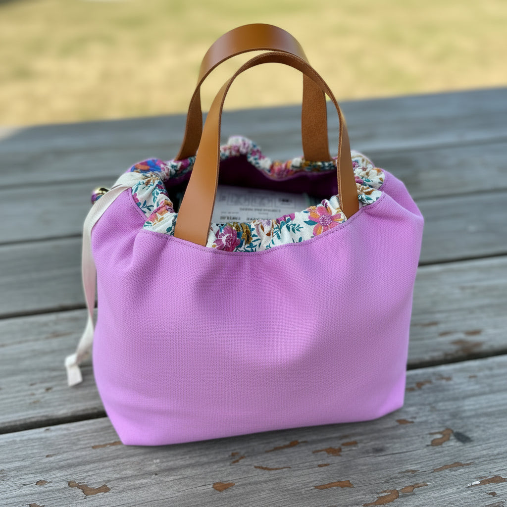 A lavender purple canvas drawstring bag with brown leather handles, a colorful floral patterned lining, and a cream drawstring resting on a white fabric background.