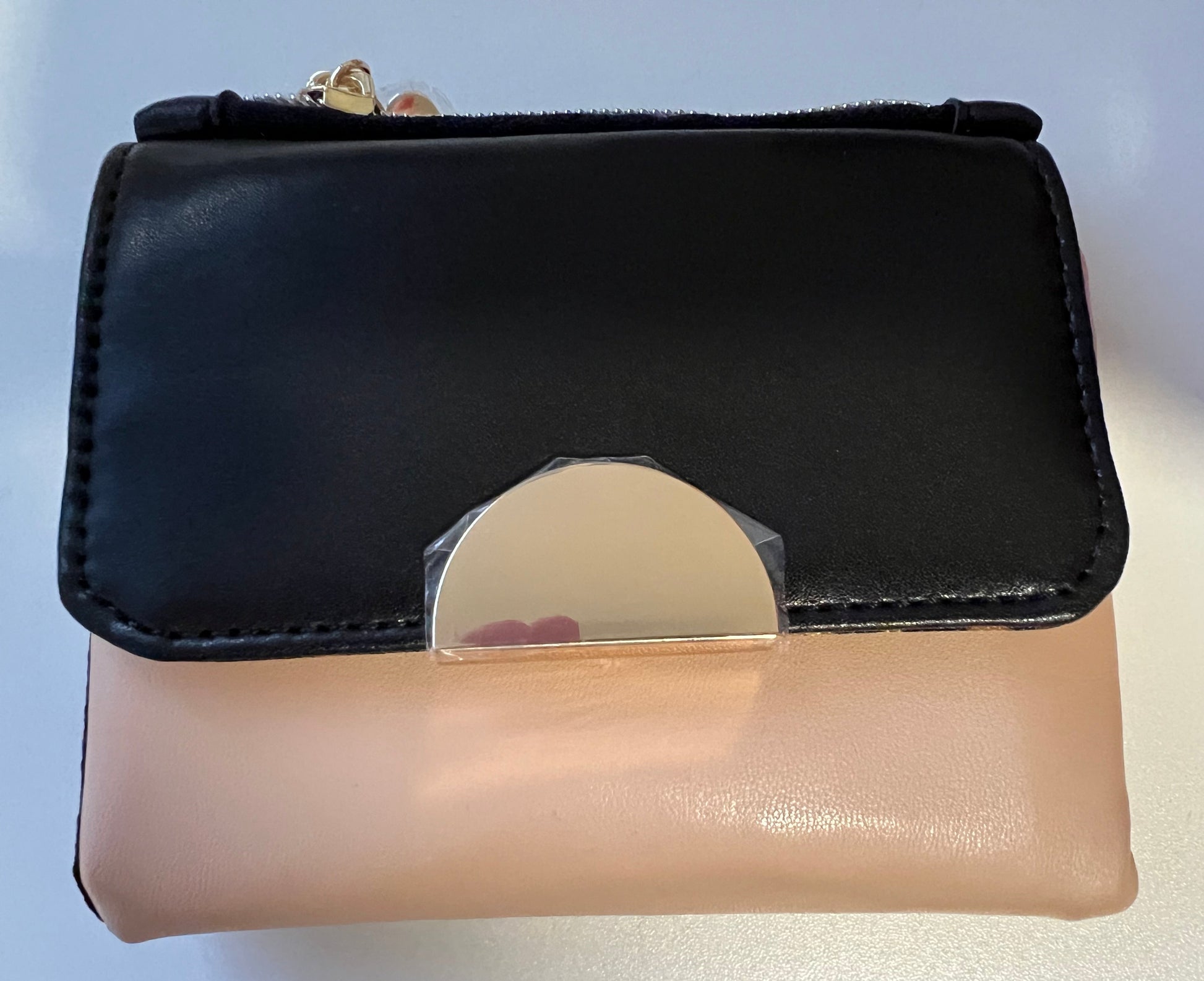 A small zip-around wallet with a black front flap and a blush pink body, featuring a large metallic gold semicircle clasp and a gold zipper pull.