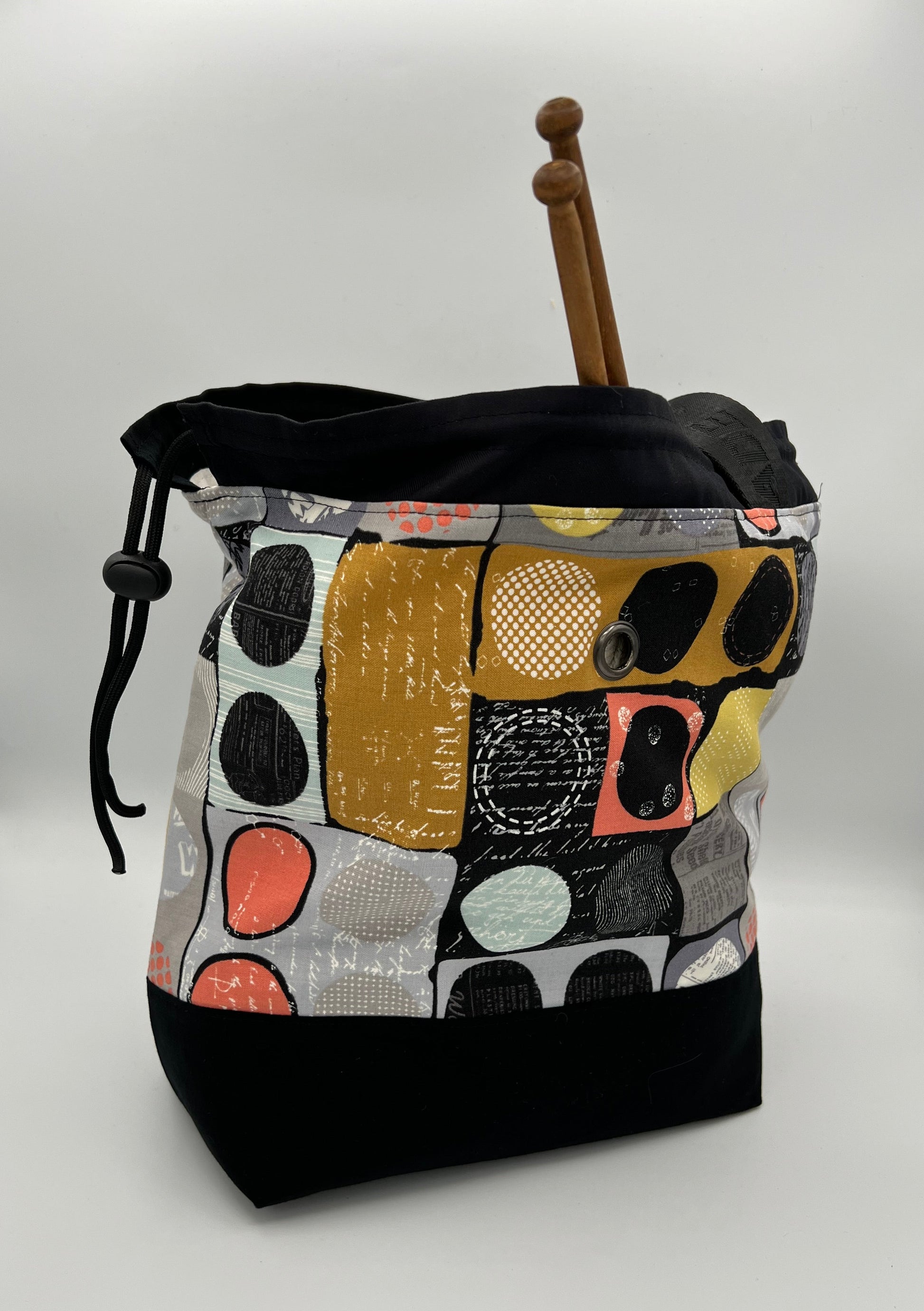 A handmade knitting bag with a black bottom and a colorful abstract patchwork pattern on the upper half. A wooden double-pointed knitting needle rests inside, and a black drawstring is visible on the left side.