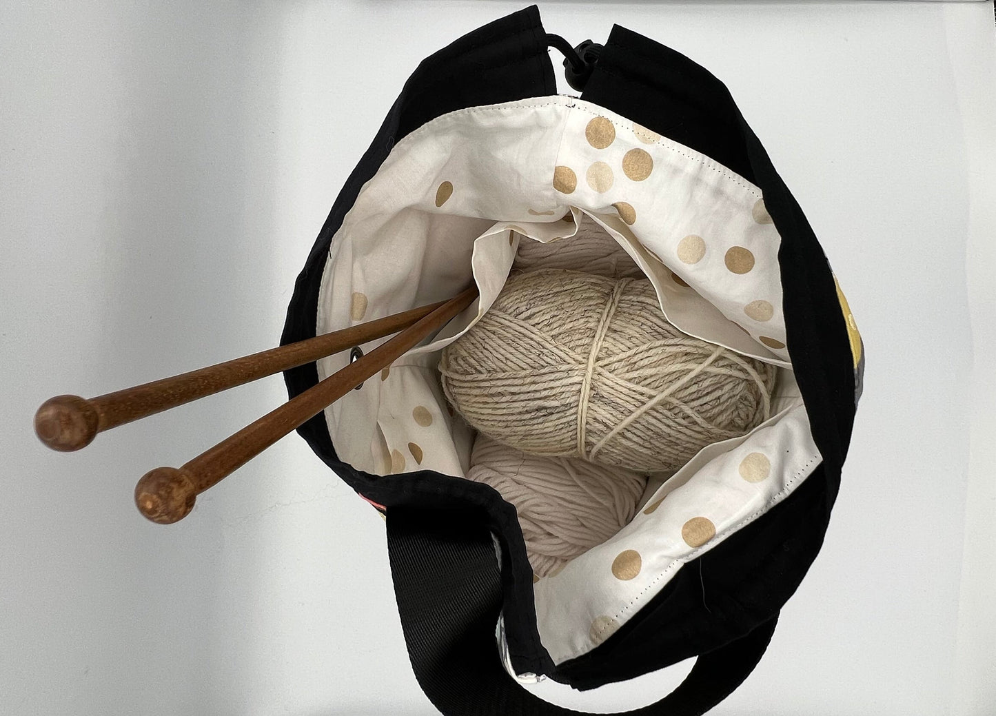 A black fabric bag with a white polka-dot interior. Inside the bag are wooden knitting needles and a ball of cream-colored yarn.
