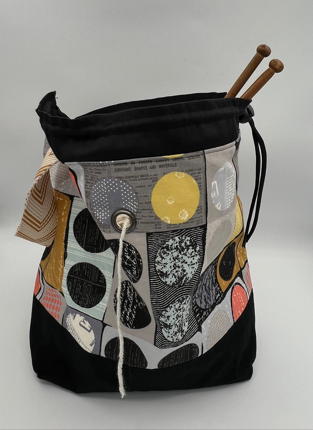 A black drawstring bag with a colorful abstract patchwork front panel featuring circles and geometric patterns in gray, yellow, white, and pink. Two wooden knitting needles are inserted into the top opening.