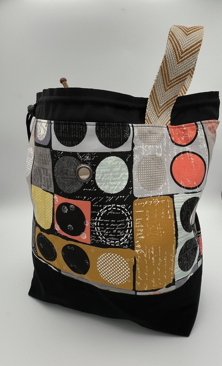 A black drawstring bag with a colorful abstract patchwork front panel featuring circles and geometric patterns in gray, yellow, white, and pink.  It has a single wide handle with a beige and brown chevron pattern.