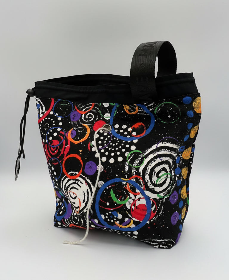 A black drawstring bag with a colorful abstract swirl pattern, featuring a black top handle and a white drawstring, photographed inside a studio.