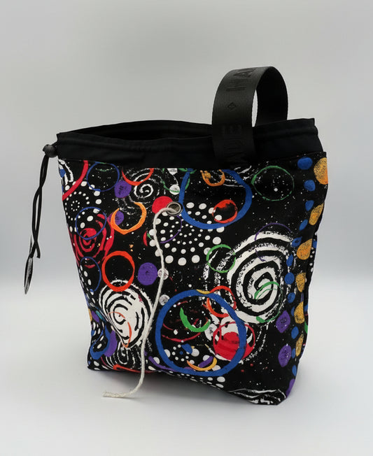A black drawstring bag with a colorful abstract swirl pattern, featuring a black top handle and a white drawstring, photographed inside a studio.
