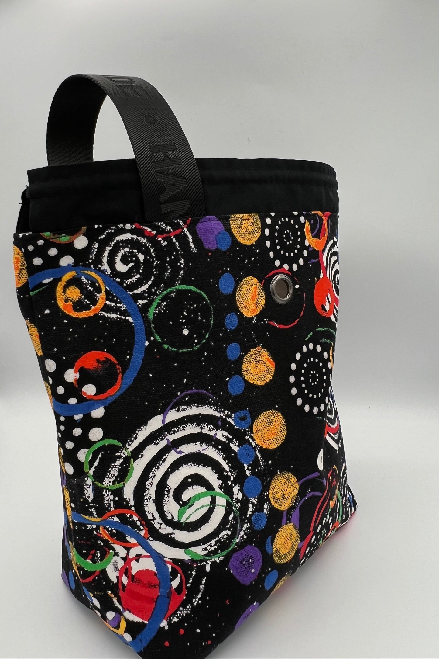 A black fabric tote bag with a colorful cosmic pattern of spirals, dots, and planets, featuring a black top handle and a snap button closure, photographed on a white studio background.