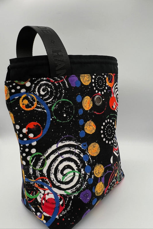 A black fabric tote bag with a colorful cosmic pattern of spirals, dots, and planets, featuring a black top handle and a snap button closure, photographed on a white studio background.