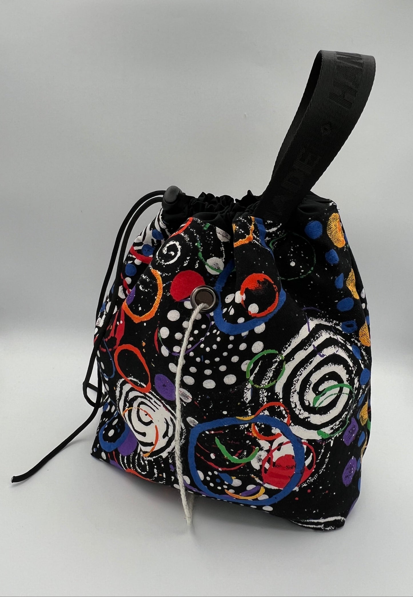 A black drawstring bag with a colorful abstract pattern of circles, spirals, and dots, featuring a black top handle and drawstring closure, photographed against a white studio backdrop.