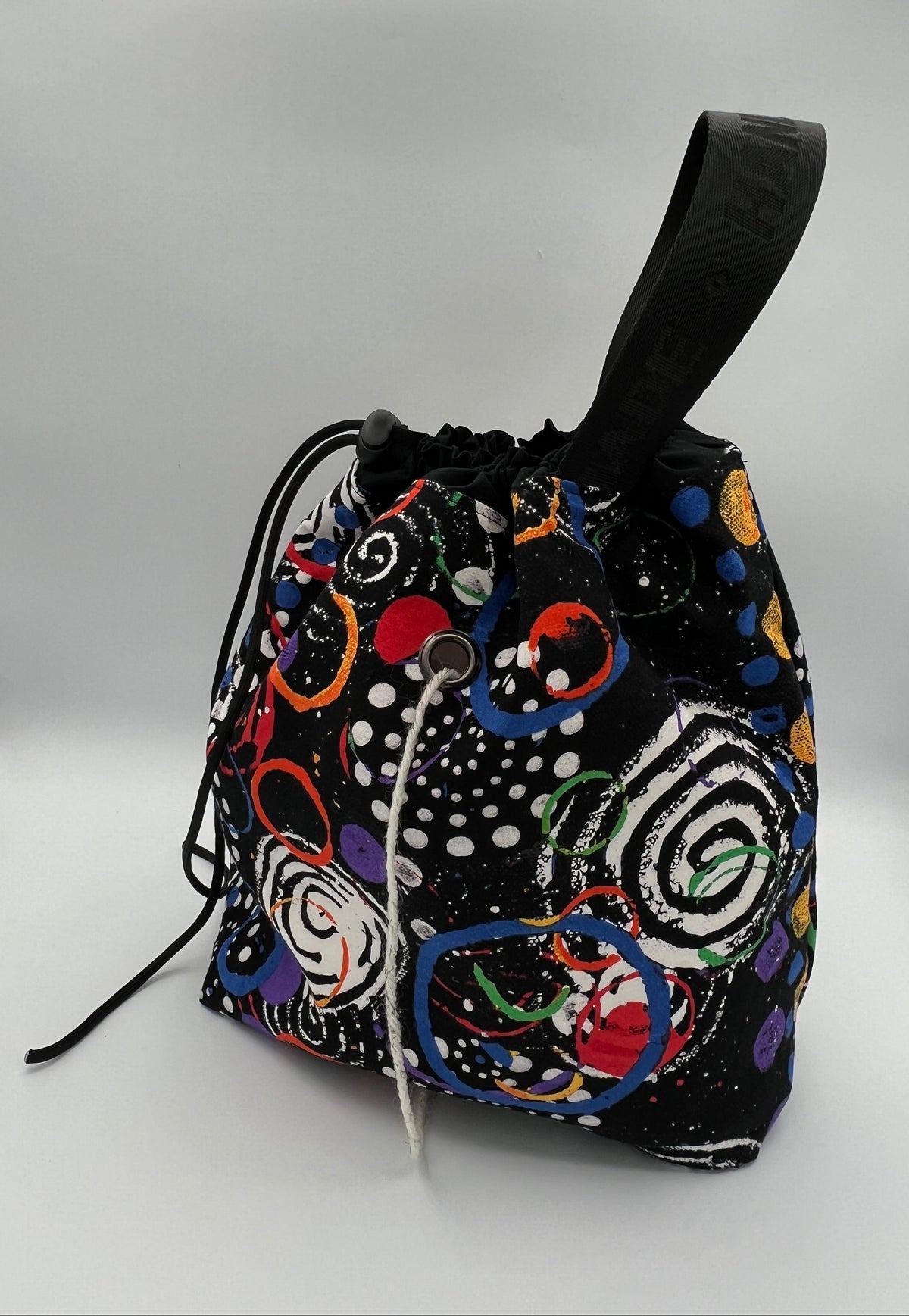 A black drawstring bag with a colorful abstract pattern of circles, spirals, and dots, featuring a black top handle and drawstring closure, photographed against a white studio backdrop.