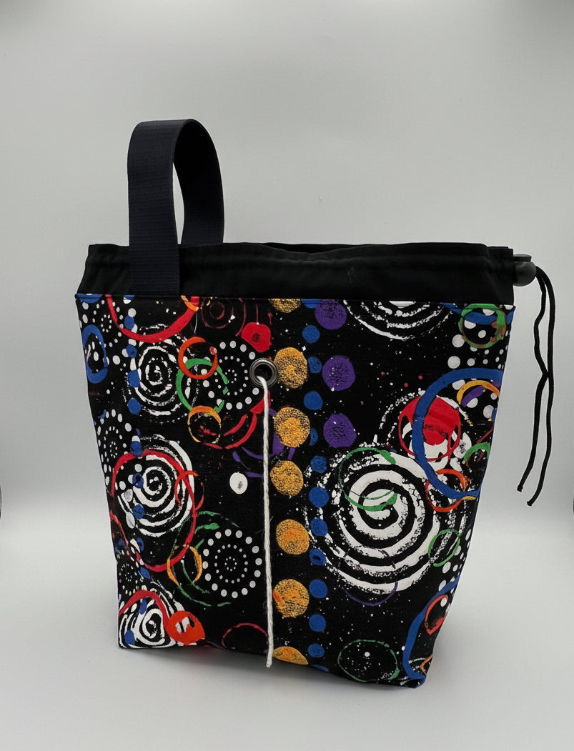 A fabric drawstring bag with a black base and a vibrant, multicolored abstract pattern of spirals, dots, and circles. The bag features a black top handle and is photographed inside a studio.