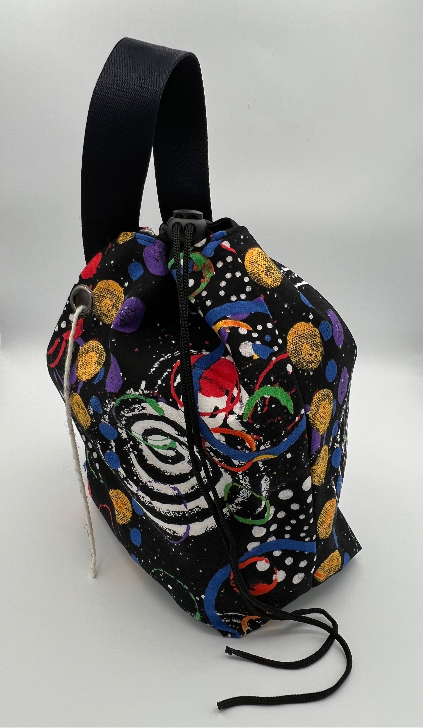 A black drawstring bag with a colorful cosmic print featuring planets, stars, and a spiral galaxy design, complete with a wide black strap, set against a white studio background.