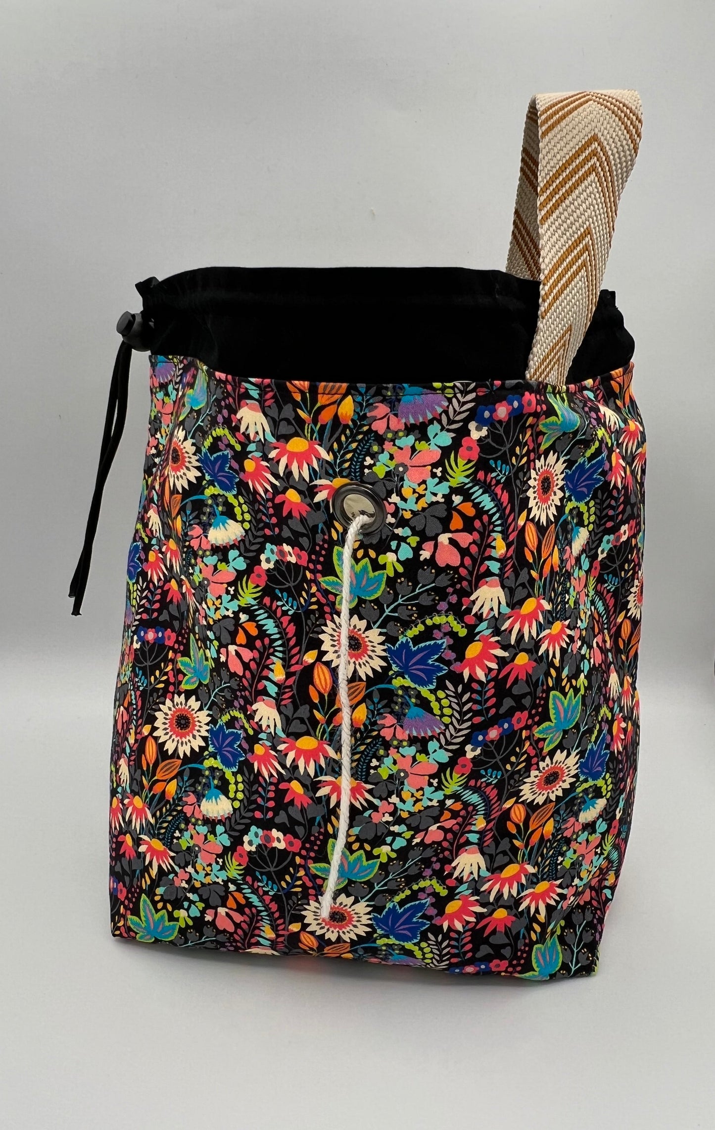 A drawstring tote bag with a vibrant multicolor floral print on a black background, featuring a black drawstring top and a beige-and-brown patterned strap, set against a plain light gray background.
