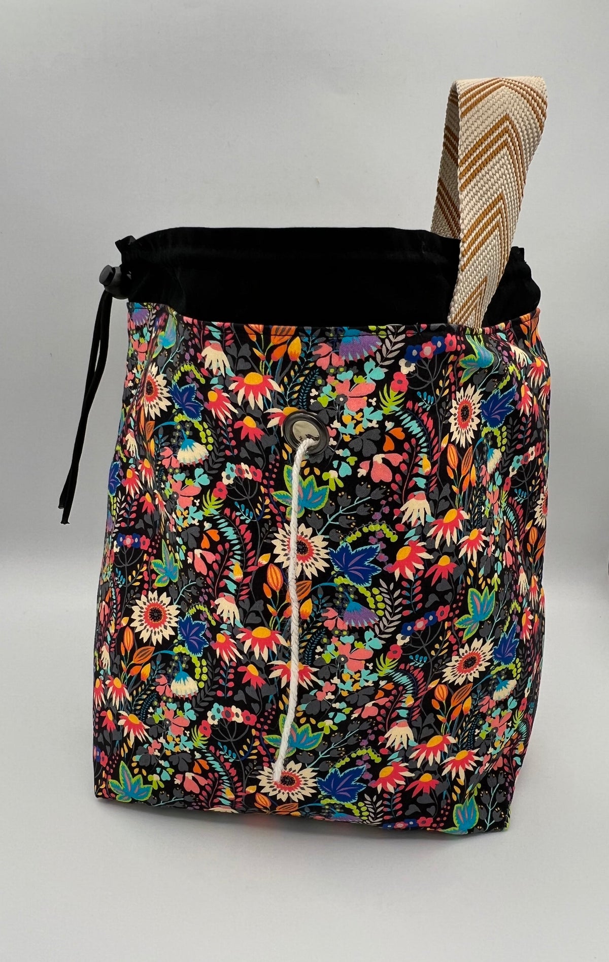A drawstring tote bag with a vibrant multicolor floral print on a black background, featuring a black drawstring top and a beige-and-brown patterned strap, set against a plain light gray background.