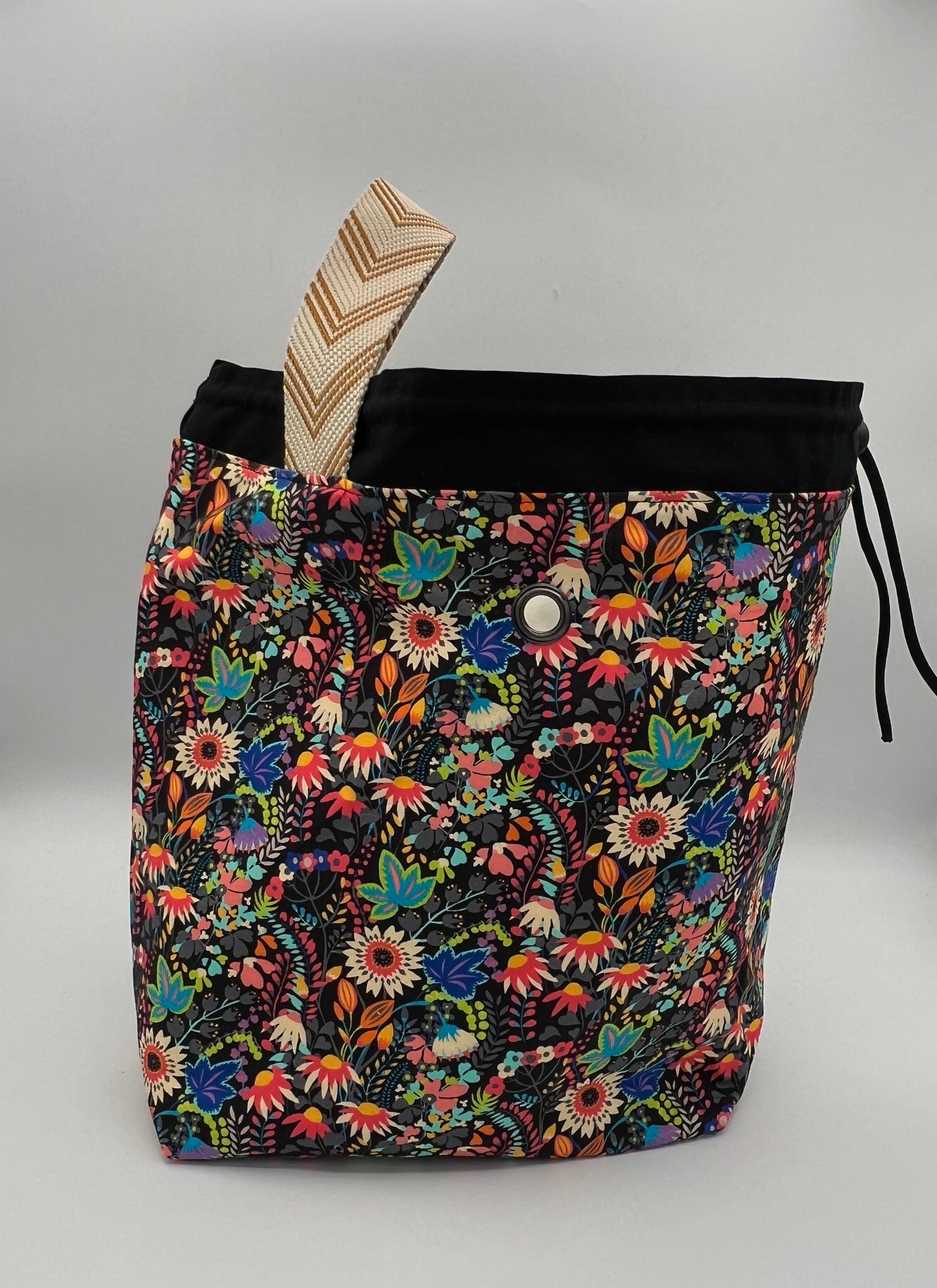 A drawstring tote bag with a vibrant multicolor floral print on a black background, featuring a black drawstring top and a beige-and-brown patterned strap, set against a plain light gray background.