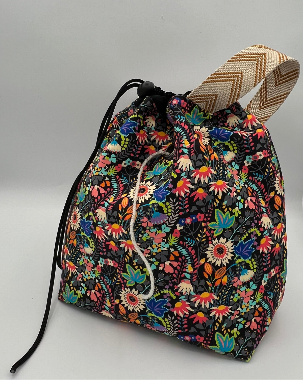 A drawstring tote bag with a vibrant multicolor floral print on a black background, featuring a black drawstring top and a beige-and-brown patterned strap, set against a plain light gray background.