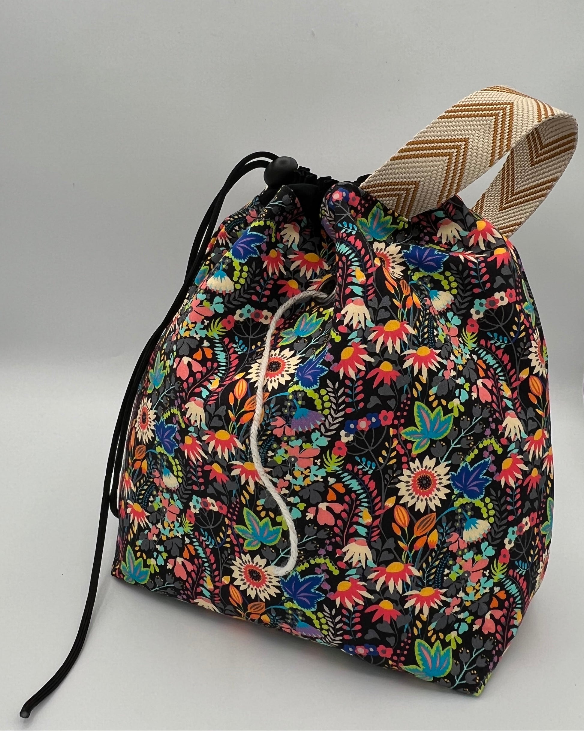 A drawstring tote bag with a vibrant multicolor floral print on a black background, featuring a black drawstring top and a beige-and-brown patterned strap, set against a plain light gray background.