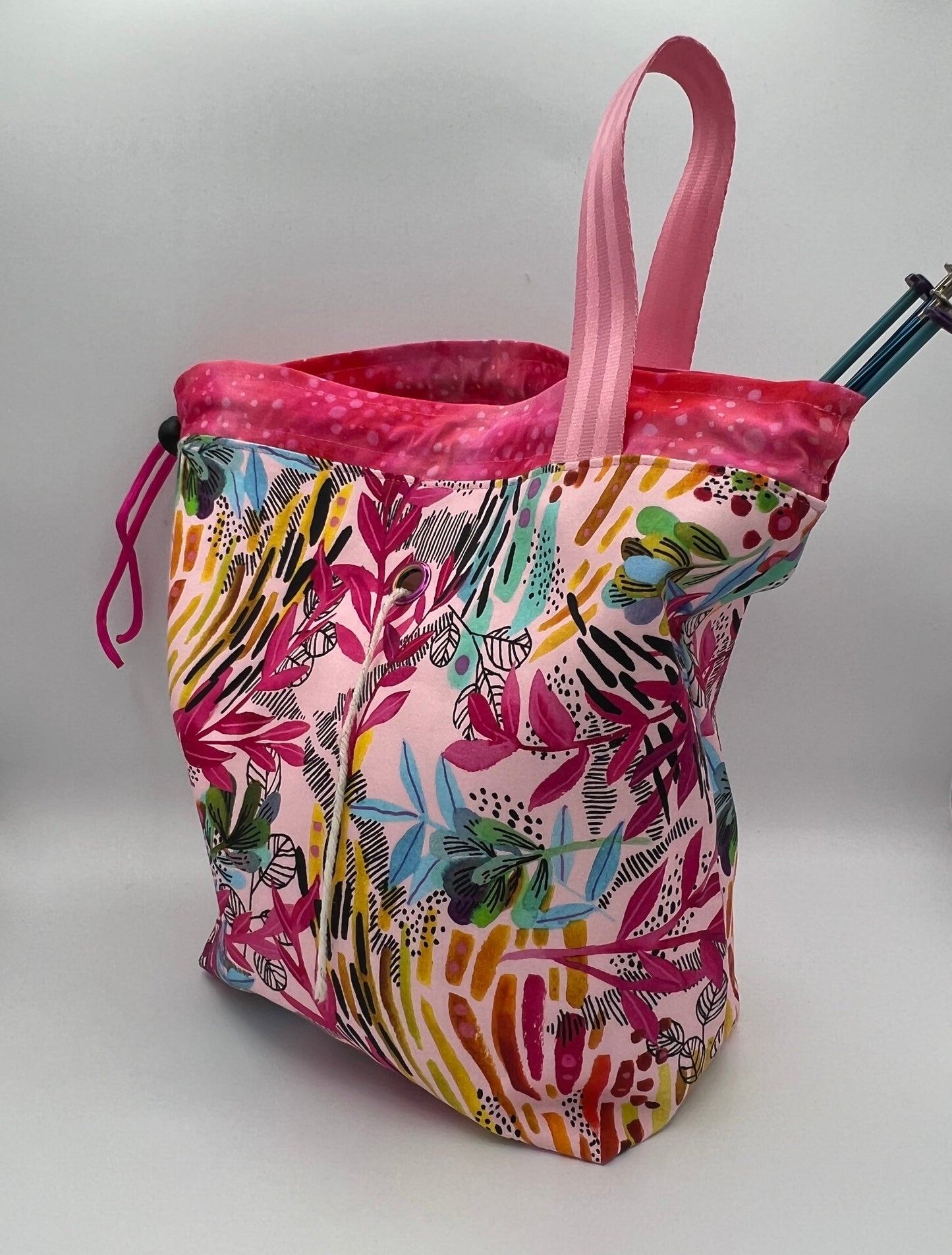 A knitting bag with a pink and multicolor botanical pattern, featuring a pink strap and a drawstring closure, photographed on a white studio background.