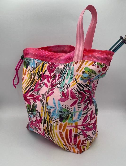 A knitting bag with a pink and multicolor botanical pattern, featuring a pink strap and a drawstring closure, photographed on a white studio background.
