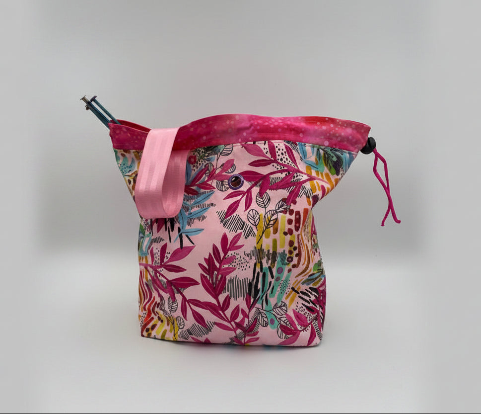 A knitting bag with a pink and multicolor botanical pattern, featuring a pink strap and a drawstring closure, photographed on a white studio background.