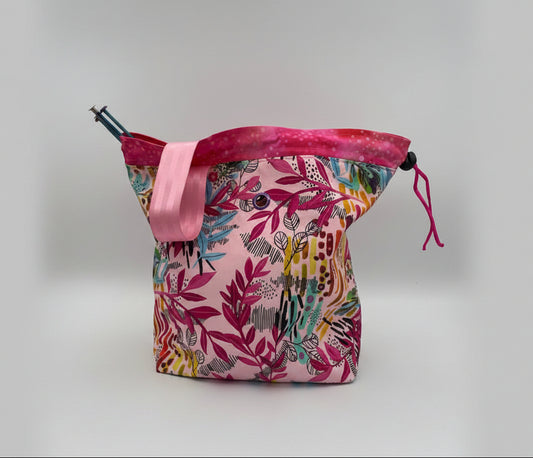 A knitting bag with a pink and multicolor botanical pattern, featuring a pink strap and a drawstring closure, photographed on a white studio background.
