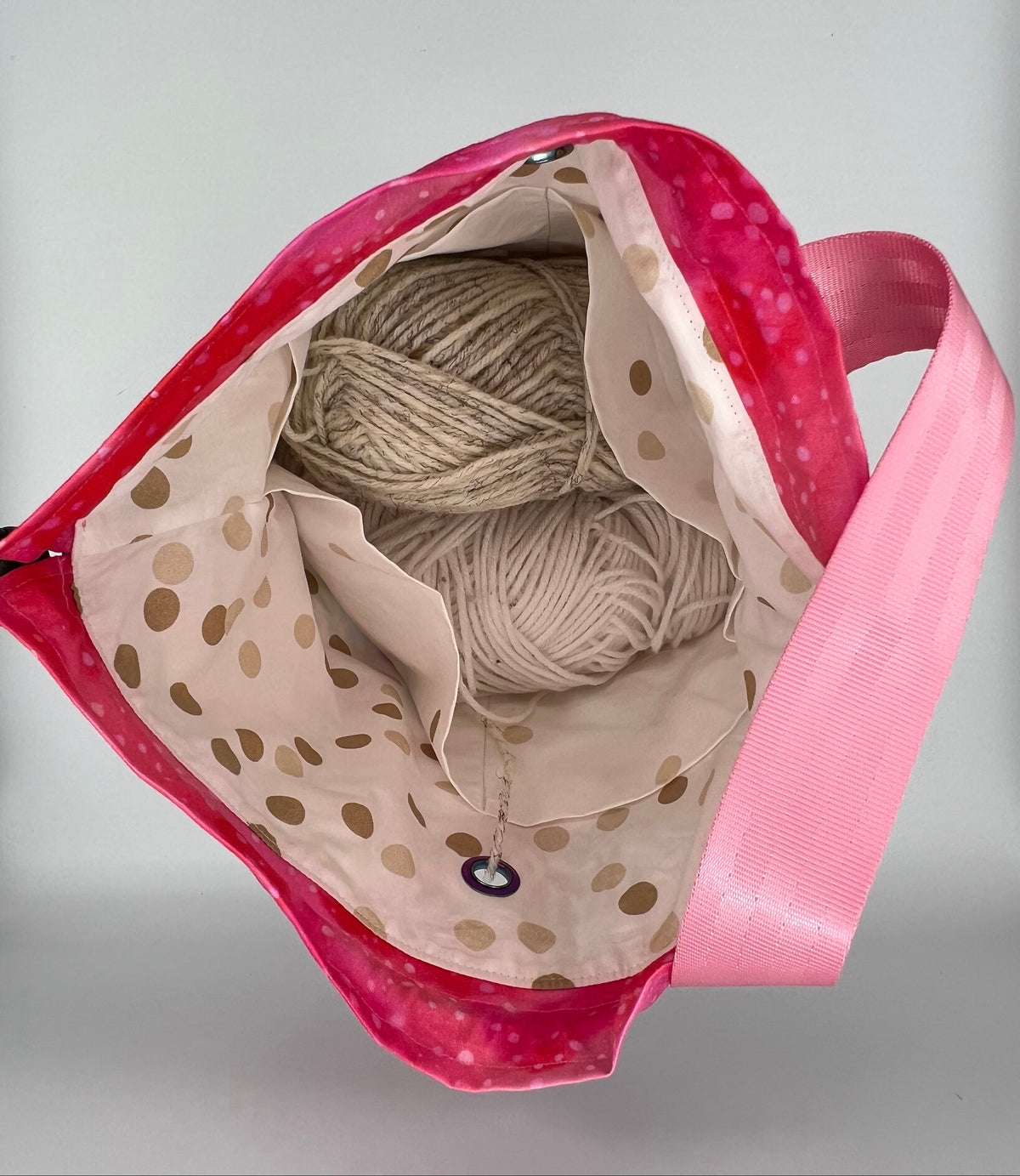 A pink fabric tote bag shown open to reveal a beige and white polka-dot lining and a pink handle. The bag is photographed against a light gray background.