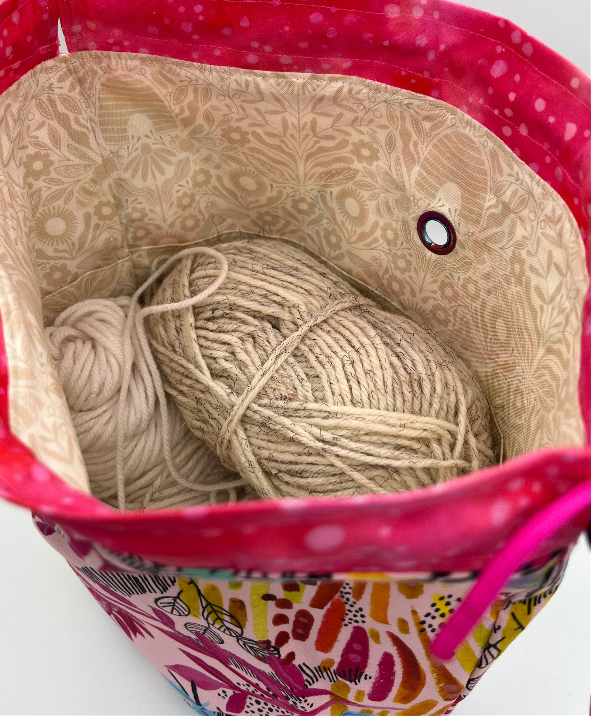 A close-up of a tote bag filled with two balls of beige yarn. The bag has a bright pink patterned rim, a beige patterned floral bee lining, and a colorful abstract print on the outside.