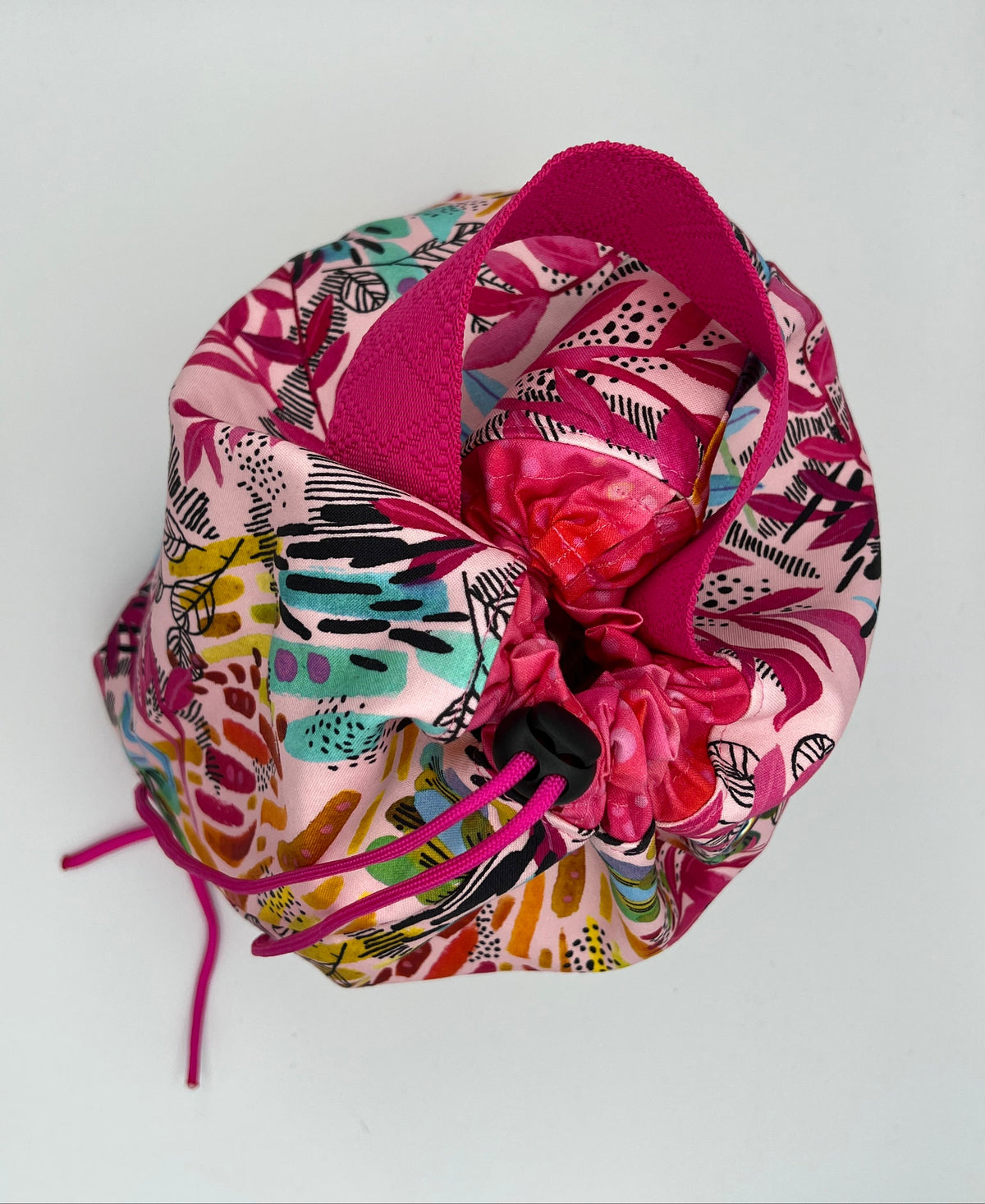A small drawstring bag with a colorful abstract print in pink, teal, yellow, orange, and black, featuring a bright pink interior lining and drawstring closure, photographed on a plain light background.