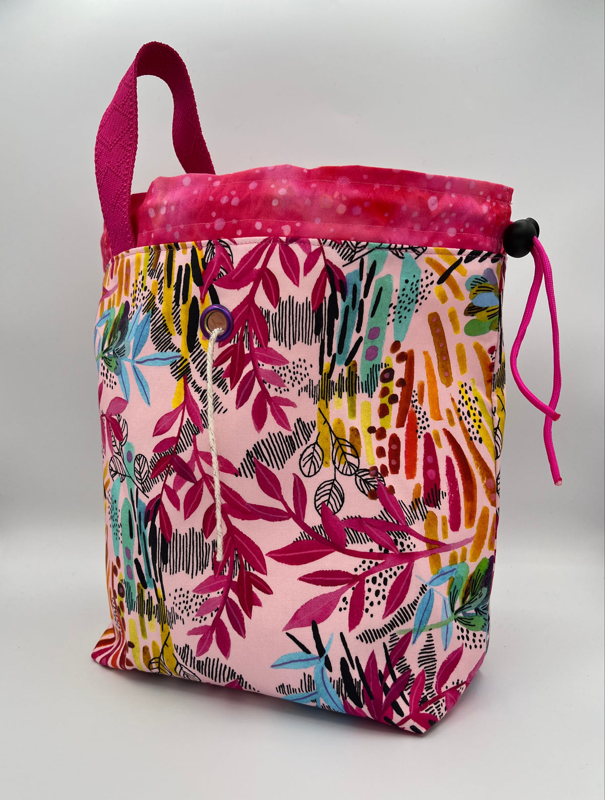 A fabric drawstring bag with a colorful tropical leaf pattern on a light pink background, featuring a solid magenta-pink top lining, top handle, and drawstring cord.