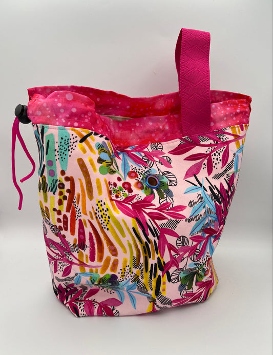 A colorful patterned fabric drawstring bag with a bright pink handle, featuring a multicolor abstract and floral print on a light pink background, set against a plain white studio backdrop.