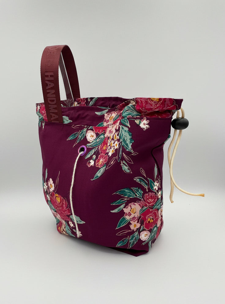A burgundy drawstring bucket bag with a floral print, featuring a burgundy strap set against a neutral studio background.