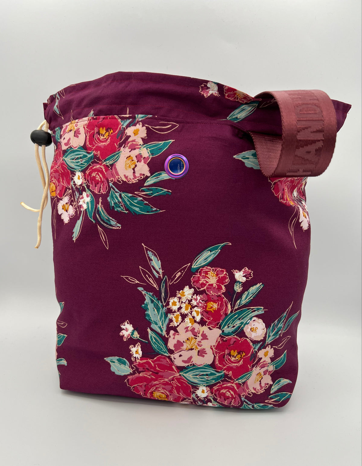 A fabric drawstring bag in deep burgundy with a colorful floral print, featuring a drawstring closure, a metal grommet, and a burgundy handle.
