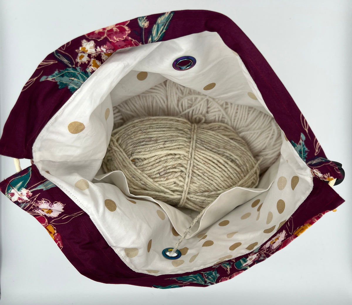 A top-down view of a fabric knitting bag, featuring a deep burgundy floral border, a white interior with gold polka dots, and a beige ball of yarn inside.