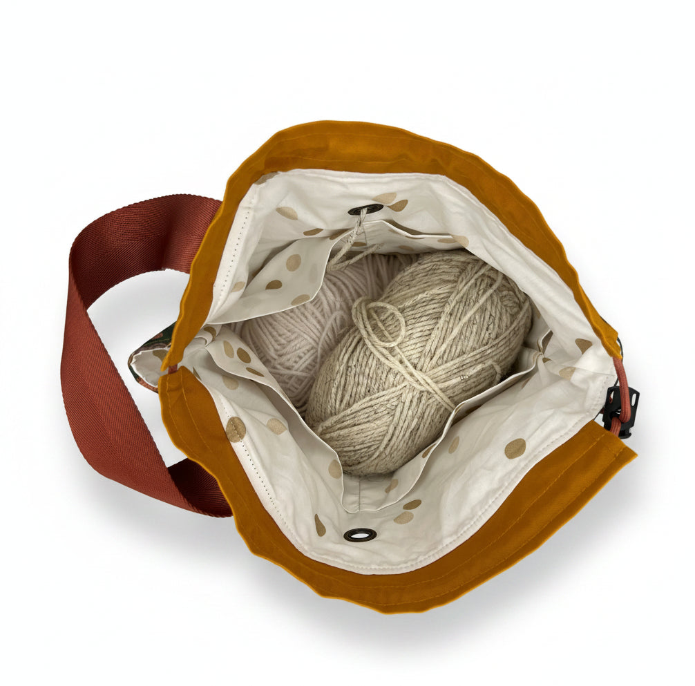 An overhead view of a mustard yellow fabric bag with a rust-colored strap, shown open to reveal a cream polka-dot interior lining holding two balls of yarn.
