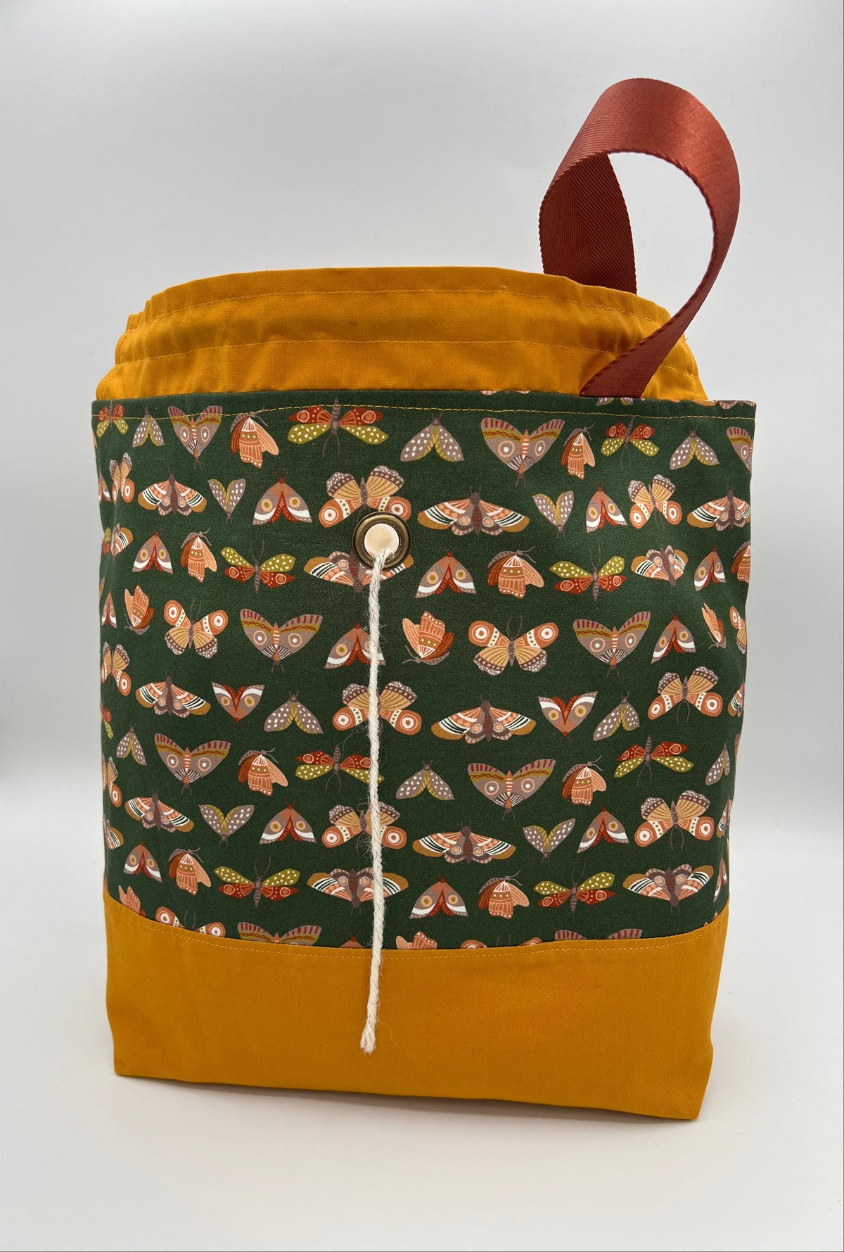 A fabric drawstring bag with a mustard yellow base and top lining, a dark green front panel covered in a repeating moth pattern, and a brown loop handle, shown on a white studio background.