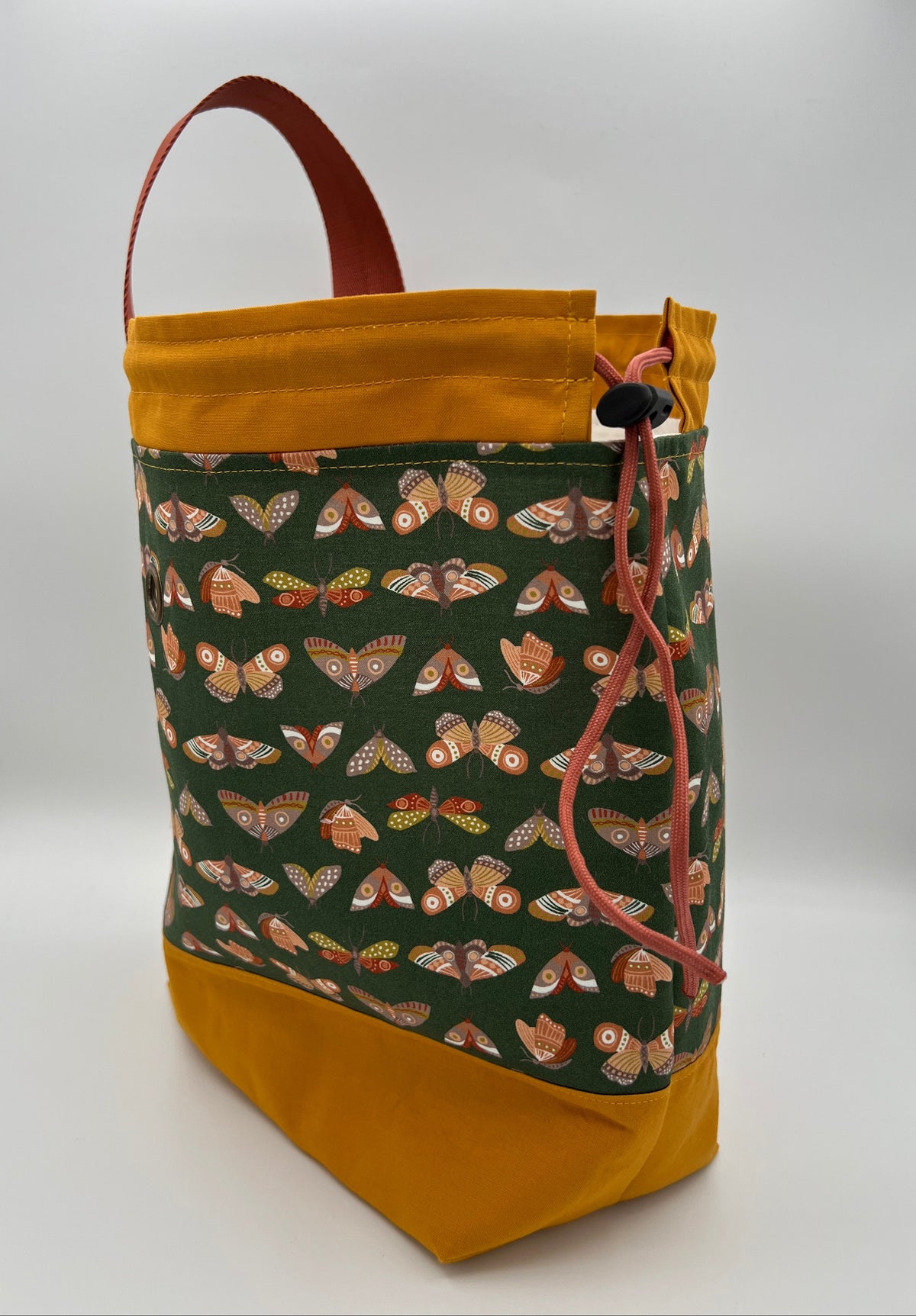 A fabric drawstring bag with a mustard yellow base and top trim, a dark green front panel covered in a pattern of orange and brown moths, and a brown top handle, shown on a white studio background.
