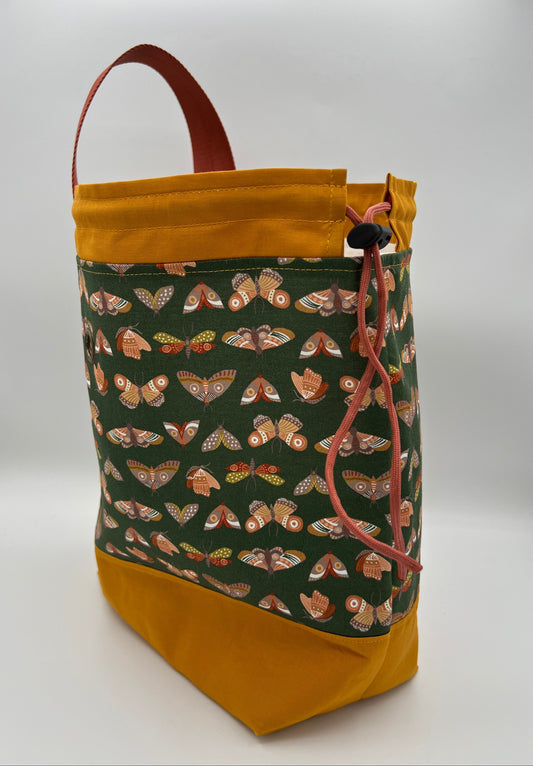 A fabric drawstring bag with a mustard yellow base and top trim, a dark green front panel covered in a pattern of orange and brown moths, and a brown top handle, shown on a white studio background.