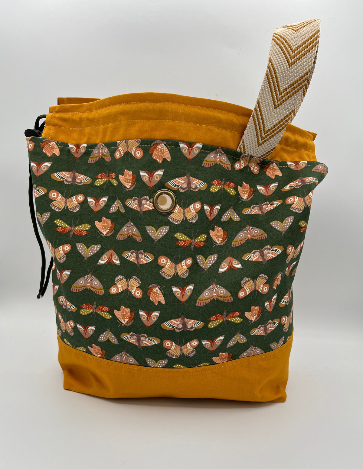 A fabric bucket bag with a dark green front panel covered in a butterfly and moth pattern, mustard yellow fabric trim at the top and bottom, a matching drawstring closure, and a woven top handle.