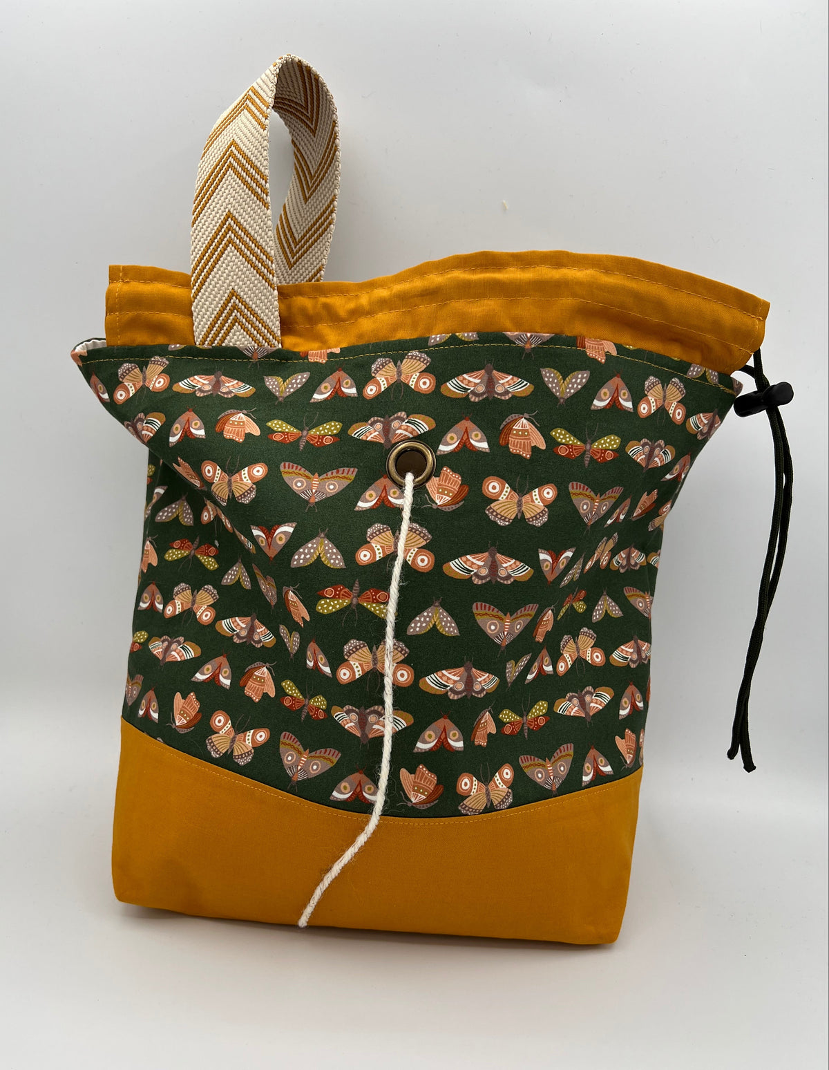 A fabric drawstring bag with a mustard yellow bottom and top trim, a dark green front panel covered in a butterfly print, and a beige-and-brown chevron-patterned top handle, shown on a light gray background.