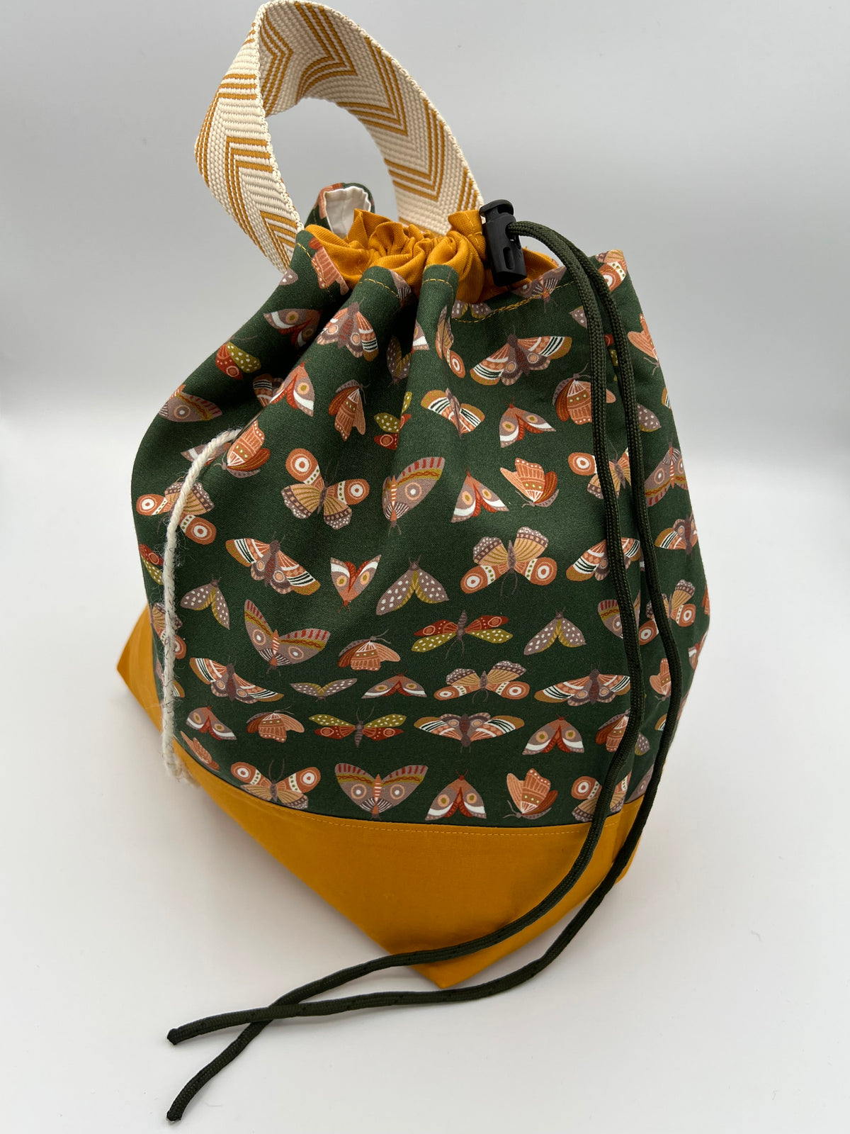 A fabric drawstring pouch with a dark green front panel printed with orange and brown moths, a mustard yellow bottom, and a striped beige-and-brown top handle, shown on a light gray background.