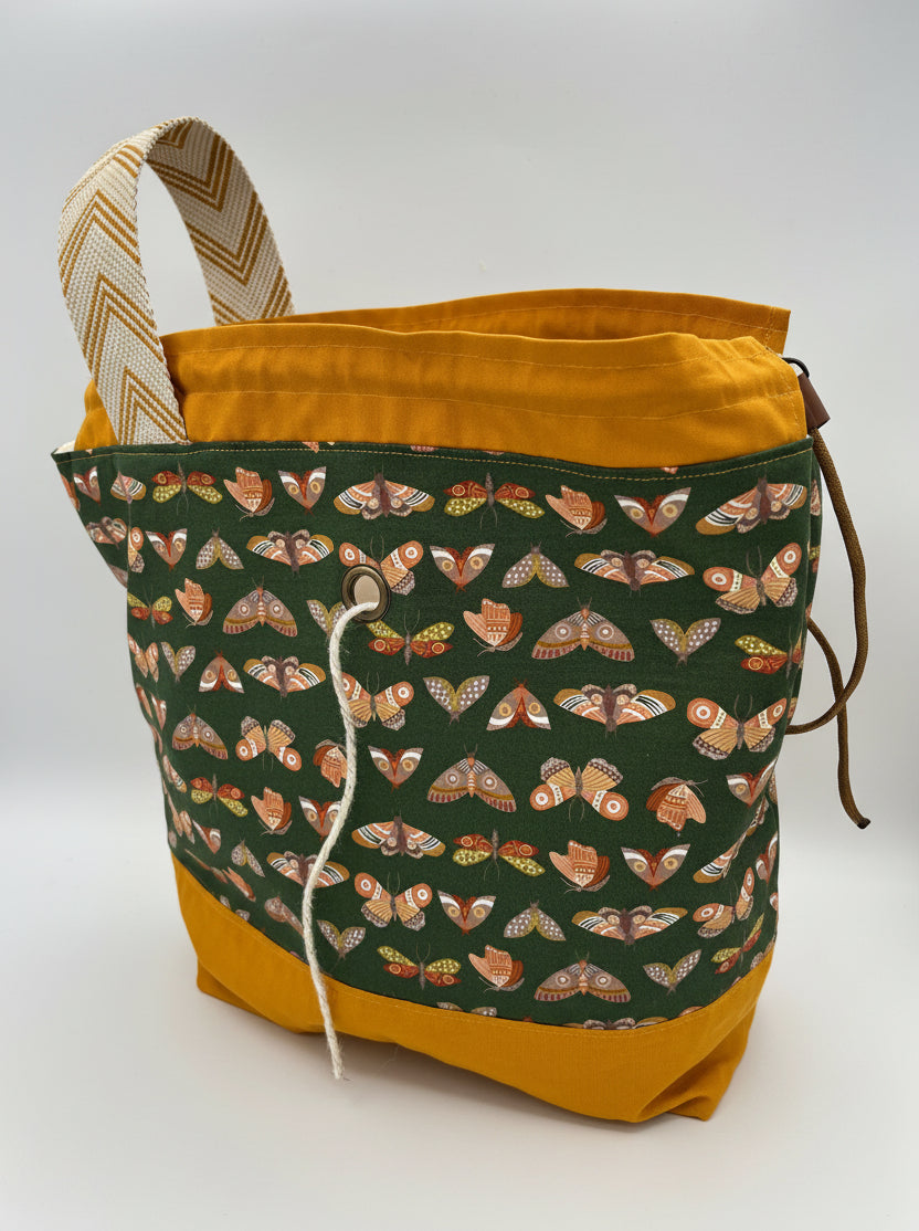 A fabric drawstring bag with a mustard yellow base and top lining, a patterned front panel featuring moths on a dark green background, and a beige-and-brown chevron-patterned handle, shown on a white studio background.