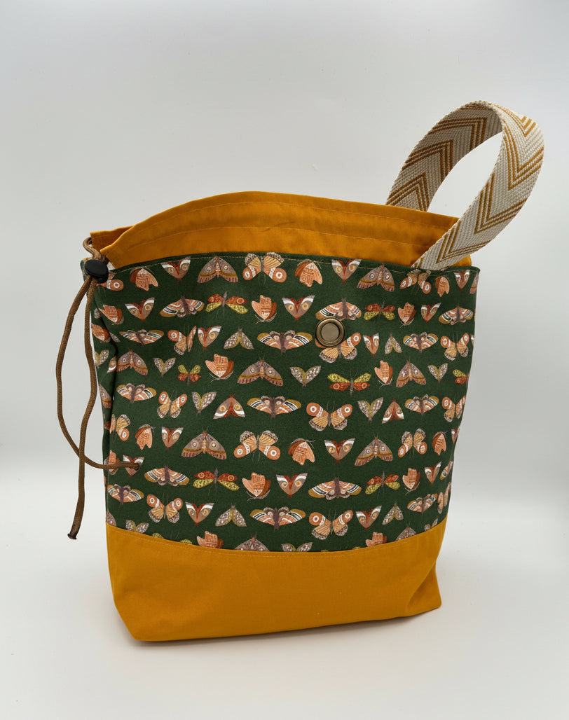 A fabric drawstring bag with a mustard yellow base and top lining, a patterned front panel featuring moths on a dark green background, and a beige-and-brown chevron-patterned handle, shown on a white studio background.
