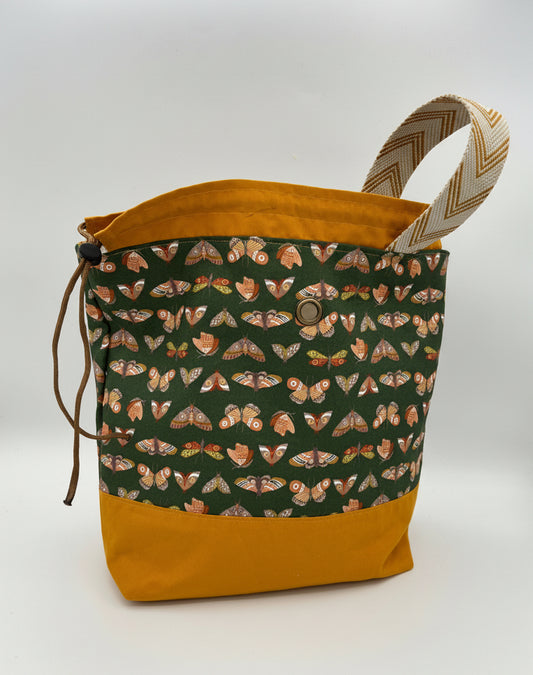 A fabric drawstring bag with a mustard yellow base and top lining, a patterned front panel featuring moths on a dark green background, and a beige-and-brown chevron-patterned handle, shown on a white studio background.
