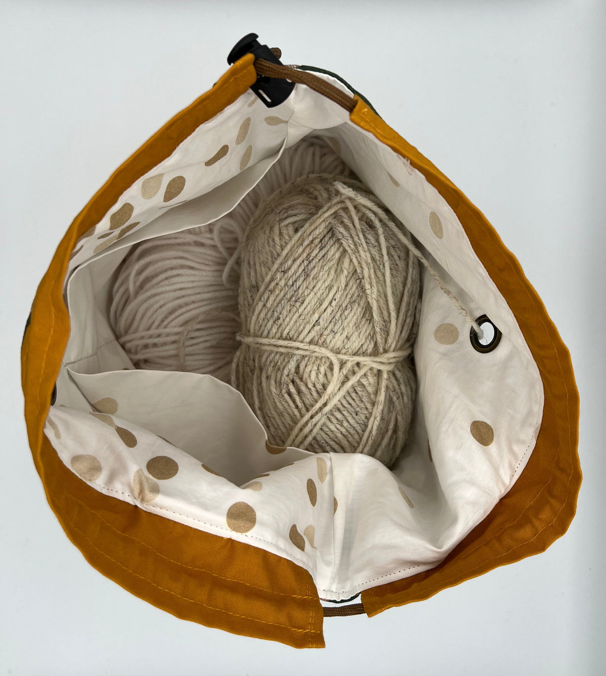 A mustard yellow fabric drawstring bag with a white polka-dot interior, shown from above containing two balls of cream-colored yarn on a white background.